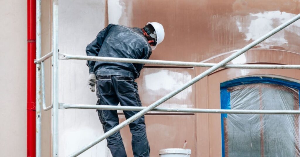 Un ouvrier refait la peinture d'une façade avec de la peinture blanche, depuis un échafaudage. 